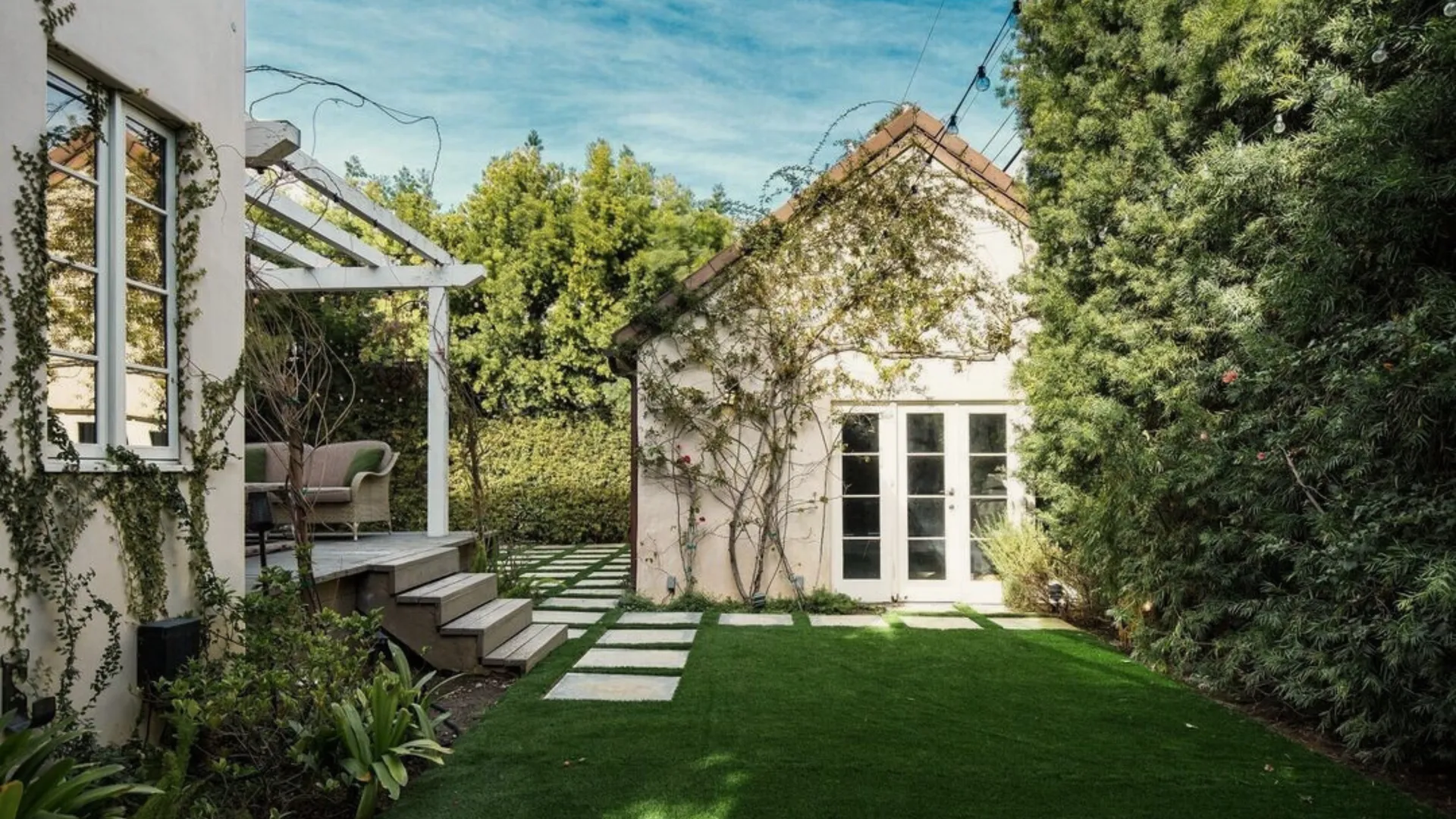 Backyard lawn with paver stepping stones leading to a detached white stucco structure with French doors, a white pergola with wicker seating off the main house and a tall hedge along the side, a Master Construction project